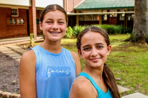 Two girls smiling outdoors; one with braces wearing a "Focus" shirt, the other with long hair. Background features a building and greenery. Youthful and cheerful.