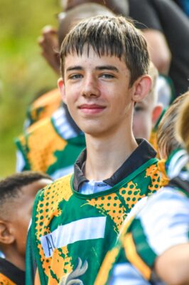 Teen boy in colorful sports uniform smiles confidently outdoors, surrounded by teammates. The mood is positive and energetic, suggesting teamwork.