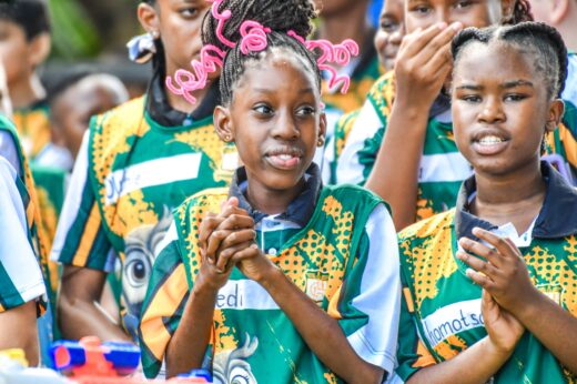 A group of children in colorful jerseys claps and cheers. They appear excited and joyful, with one girl notably sporting bright pink hair accessories.