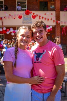 A smiling couple in pink shirts embraces in front of a decorated building with hearts and balloons, creating a joyful, festive atmosphere.