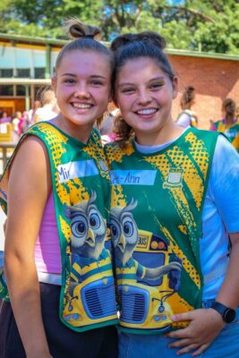 Two smiling girls wearing colorful vests with owl and school bus graphics stand closely together outdoors on a sunny day, with trees and a building in the background.