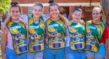 Five smiling girls with top-knot hairstyles pose closely, wearing green shirts with cartoon owls, set against a sunny schoolyard. The mood is cheerful.