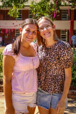Two smiling women stand outdoors in a sunny courtyard with green trees. One wears a pink blouse, the other a leopard print top and glasses.