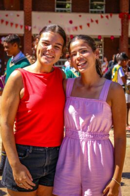 Two women smiling warmly, one in a red top and black shorts, the other in a pink dress. They stand outdoors at a festive event with colorful decorations.