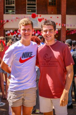 Two young men smile outdoors at a festive event, with heart decorations and people in the background. One wears a white Nike t-shirt, the other an Old Khaki shirt.