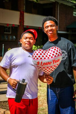 Two people smiling outdoors. One holds a heart-shaped balloon with "I Love You" text, while the other holds a black tumbler. A bright, cheerful atmosphere.
