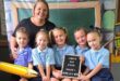 A teacher and five young students in blue uniforms sit together smiling. Behind them is a colorful backdrop with hearts and a chalkboard. In front, there’s a globe, paintbrushes, and an abacus. The children hold a sign with text in another language. The scene conveys a cheerful and welcoming back-to-school atmosphere.