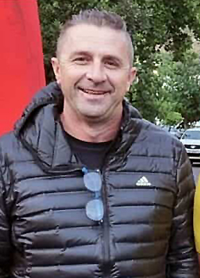 A man in a black puffer jacket smiles while standing outdoors. Sunglasses hang from his jacket. Trees and a red flag are in the background, conveying a casual, cheerful atmosphere.