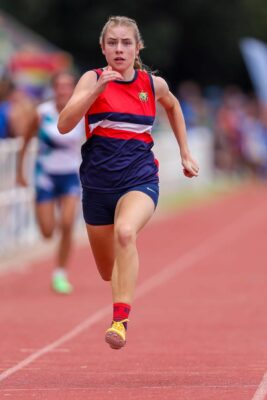 A determined female athlete in a red and blue uniform sprints on a track, focused and intense. It's a bright day, and blurred competitors trail behind.