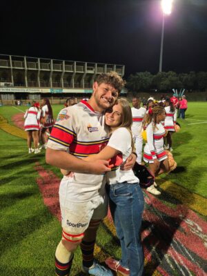 A rugby player in a uniform embraces a smiling woman on a floodlit field. Cheerleaders in matching outfits are visible in the background, creating a celebratory mood.