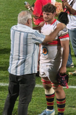 An older man comforts a rugby player on the field. The player wears a white jersey and holds a helmet, appearing emotional. Supportive atmosphere.