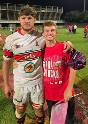 Two young men stand on a rugby field at night, smiling. The left one wears a white rugby uniform; the right one wears a red T-shirt.