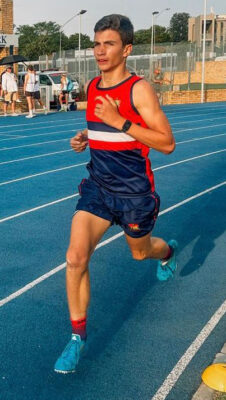 Young athlete in a blue and red uniform runs confidently on a blue track under clear skies, conveying determination and focus. People and a car are visible in the background.