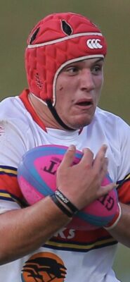 Rugby player wearing a red helmet and white jersey grips a pink and blue ball, looking focused and determined. The scene conveys action and intensity.