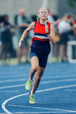 A female athlete in a red and blue uniform and lime green shoes sprints on a blue track, focused and determined, with a blurred audience in the background.
