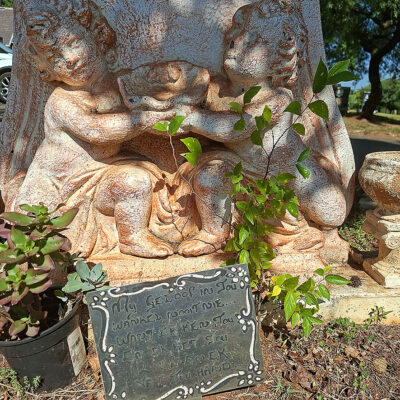 A weathered stone sculpture of two cherubs playfully exchanging a bowl is surrounded by green plants. A small chalkboard sign adds a whimsical touch.