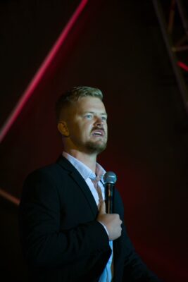 Man in a suit passionately speaking into a microphone on stage, with dramatic lighting and a dark background, conveying intensity and focus.