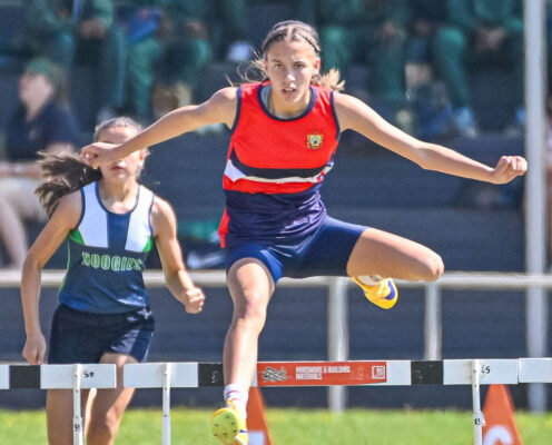 A young athlete in a red and blue uniform leaps over a hurdle with focus and determination during a race, while another competitor follows closely behind.
