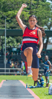 Athlete in red top, blue shorts mid-jump on a long jump track; arm raised, focused expression. Sunny day, trees and spectators in background.