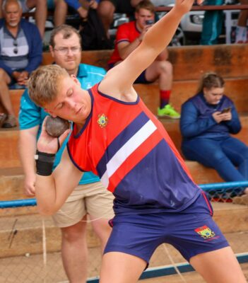 Young athlete in red and blue uniform prepares to throw a shot put, with spectators blurred in the background. Focused expression, outdoor setting.