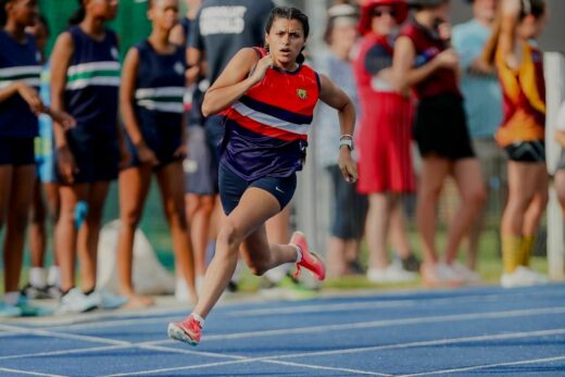 A female athlete sprints on a blue track, wearing a red and navy uniform with white stripes. Focused expression. Spectators stand in the background.