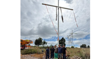 A group of five utility workers stand in front of a tall metal pole with crossbars on a cloudy day. Nearby are a crane and electrical towers. The mood is collaborative.