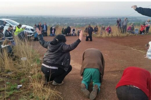 A group of people kneel and pray on a dirt road with hands raised, surrounded by dry grass and cars. The scene conveys a sense of devotion and unity.