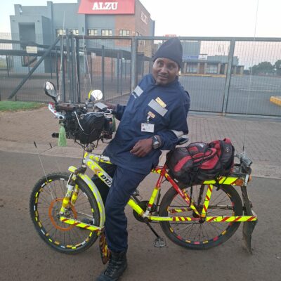 A person in a navy uniform leans against a brightly decorated bicycle with reflectors and a red backpack. They smile, conveying a relaxed, cheerful mood.