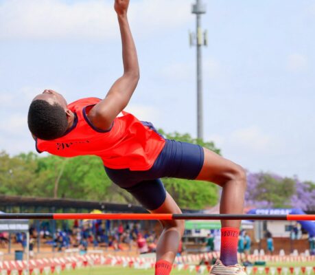 Athlete in a red uniform gracefully clears a high jump bar against a blue sky, showcasing focus and athleticism. Vibrant outdoor sports setting.