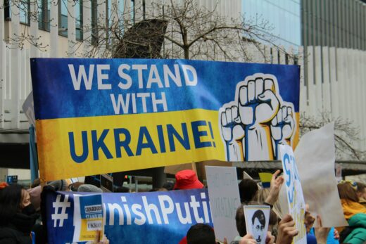 Protesters hold a banner reading "We Stand With Ukraine" against a blue and yellow background. Raised fists symbolize solidarity. Mood: supportive, determined.