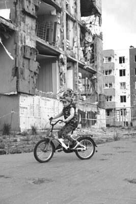 A young child rides a bicycle in front of a heavily damaged building. The scene is somber, with rubble and destruction evident in the background.