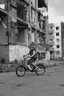 A child rides a bicycle in front of a partially destroyed building. The scene is in black and white, conveying a feeling of resilience amid devastation.