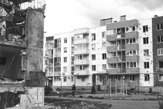 A black-and-white image shows a war-torn urban area with heavily damaged buildings. debris, shattered windows, and a somber, desolate atmosphere.