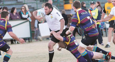A rugby player in a white jersey fiercely runs with the ball, evading tackles from opponents in striped jerseys on a grassy field. The crowd watches intently.