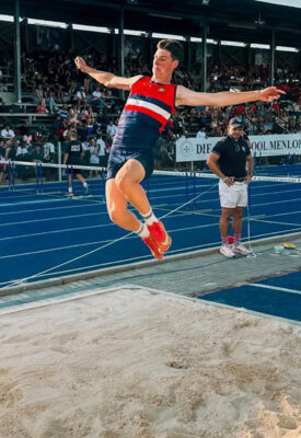Athlete in midair performing a long jump, wearing a blue and red uniform with red shoes. A sandpit and track are visible, with a crowd and official looking on.