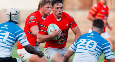 Two rugby players in red jerseys advance with the ball, focused and determined, while two opponents in blue attempt to block them on a grassy field.