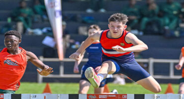 Two young male athletes in vibrant sportswear leap over hurdles on a sunny track, displaying focus and determination during a competitive race.