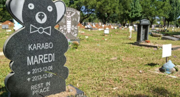 A teddy bear-shaped headstone with "Karabo Maredi" inscribed sits in a sunlit cemetery. Green grass, flowers, and other tombstones are in the background.