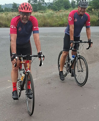 Two cyclists in red and navy jerseys and helmets ride along a road, surrounded by greenery. Both appear relaxed, enjoying an outdoor ride.