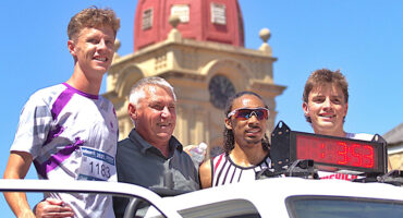 Four men pose cheerfully on a vehicle in front of a historic building with a red dome. One wears a race bib, and a timer reads "3:53." The atmosphere is celebratory.