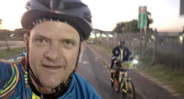 A cyclist in a helmet and colorful jersey smiles at the camera, with another cyclist in the background on a road at dusk. The mood is lively and adventurous.
