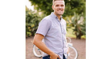 A man in a light blue shirt and jeans stands outdoors, smiling warmly. A bicycle is visible in the blurred background, with trees adding a serene feel.