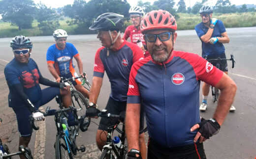 Group of seven cyclists in colorful jerseys and helmets pause on a roadside, smiling and talking, conveying a cheerful, relaxed atmosphere.