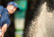 Golfer swings in a sand trap, sending a spray of sand into the air. He wears a navy cap and shirt. The scene captures focus and intensity.