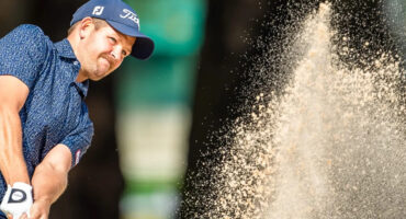 Golfer swings in a sand trap, sending a spray of sand into the air. He wears a navy cap and shirt. The scene captures focus and intensity.