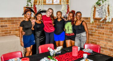Six smiling people stand together in front of a decorated brick wall. A table with red candles and floral centerpieces is in the foreground, creating a warm, celebratory atmosphere.