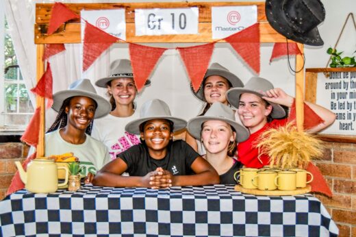 Six smiling people in hats, standing behind a checkered table adorned with mugs and a teapot. A "Gr 10" sign and festive bunting are above them.