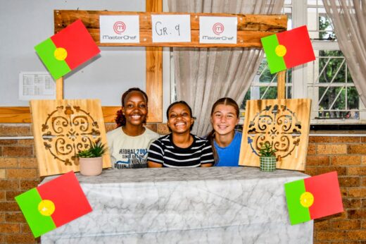 Three smiling students are behind a table with a "MasterChef Gr. 9" sign. Decorative wooden panels, colorful flags, and a small plant are displayed. Bright, cheerful atmosphere.