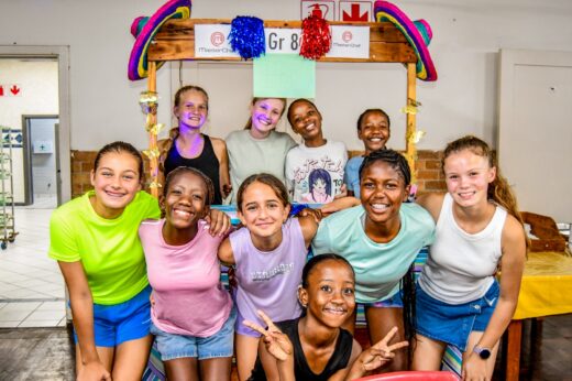 A joyful group of nine children, smiling and posing under a brightly decorated structure labeled "Gr 8." They appear happy and vibrant.