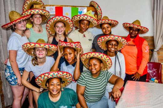 A group of smiling people wearing colorful sombreros pose indoors. A festive poster with flags is in the background, creating a joyful atmosphere.